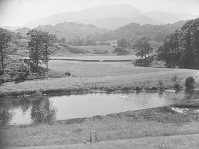 Mujeres empujando a un bebé en un cochecito en la orilla del río a través de un prado con gente nadando en el río y un agricultor trabajando con un tractor en los campos más allá y montañas al fondo. de Joseph Hardman