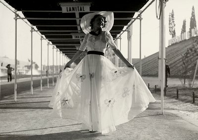 Modelo en un vestido de verano completo creado por la casa de alta costura "Graldi". El vestido es de una tela ligera decorada con bordados; un sombrero de ala ancha completa el conjunto. de Luis Bogino