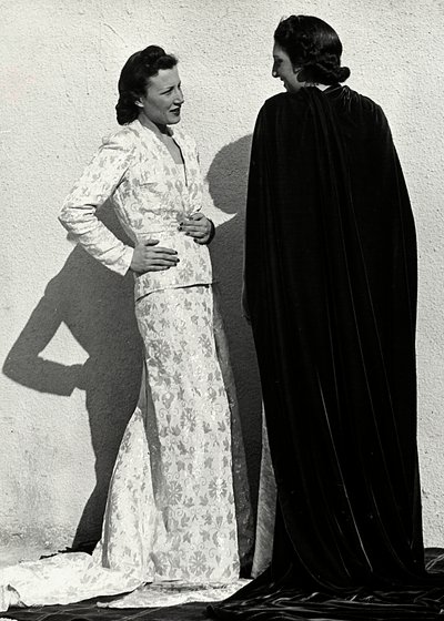 Dos modelos presentan creaciones de la casa de alta costura italiana "Dragoni". Una modelo lleva un conjunto de material estampado compuesto por una chaqueta y una falda larga con cola. La otra modelo, vista desde atrás, lleva un manto largo de terciopelo. de Luis Bogino