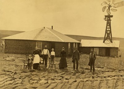 Familia de colonos, fuera de su casa de tepes con un molino de viento en el tejado del edificio contiguo en el condado de Custer, Nebraska, hacia 1885 (fotografía en blanco y negro). de Solomon D. Butcher