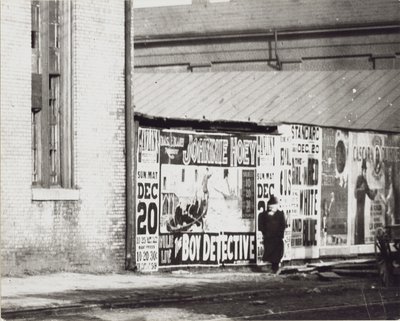 Un hombre pasa junto a una pared de anuncios de carteles, incluido uno de Johnnie Hoey, el niño detective, que se presenta en el Teatro Havlin en 600 Walnut Street. de Unbekannt