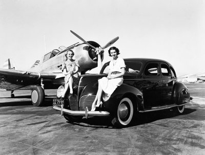 California: 1939 Dos mujeres sentadas en los guardabarros de un Lincoln Zephyr de 1939 en un aeropuerto con un avión de un solo motor estacionado detrás de ellas (foto en b/n) de Unbekannt