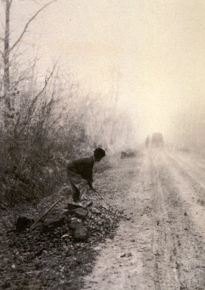 Un camino rural envuelto en la bruma matinal. En primer plano un hombre parte piedras de Unbekannter Künstler