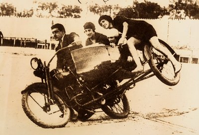 Un hombre se exhibe conduciendo una motocicleta con un sidecar en el que viajan tres mujeres jóvenes. El sidecar se levanta del suelo al tomar una curva. de Unknown photographer