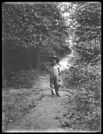 William Gray Hassler con sombrero de paja y overol, llevando una caña de pescar y una cesta, caminando por un sendero en el bosque solo de William Davis Hassler