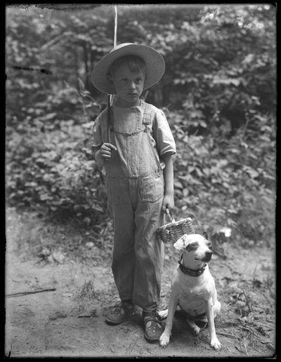 William Gray Hassler con sombrero de paja y overol, llevando una caña de pescar y una cesta, posando en el bosque con un perro pequeño (Bessie), 20 de agosto de 1911 de William Davis Hassler