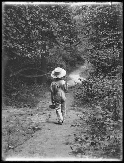 William Gray Hassler con sombrero de paja y overol, llevando una caña de pescar y una cesta, caminando por un sendero en el bosque solo, 20 de agosto de 1911 de William Davis Hassler