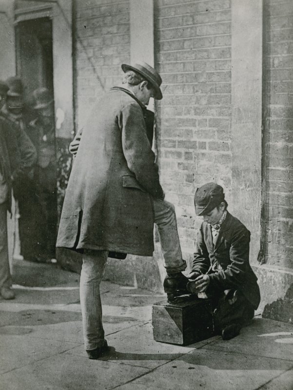Un joven limpiabotas lustrando los zapatos de un hombre en la calle de English Photographer