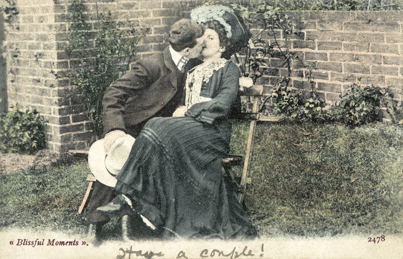 Hombre robando un beso a una chica en un banco de jardín de English Photographer