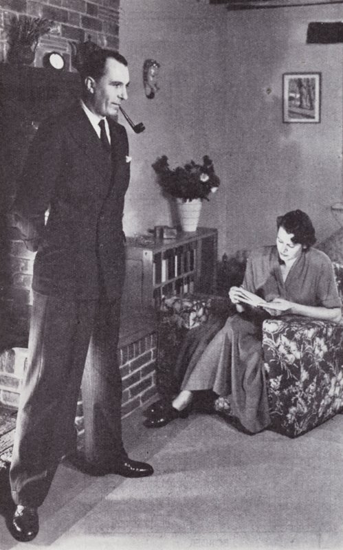 Esposa lee, mientras el esposo calienta su espalda contra el fuego, c.1950 de English Photographer
