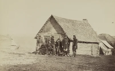 Un grupo de soldados y dos jóvenes, uno afroamericano, fuera de las cabañas de troncos, 1861-65
