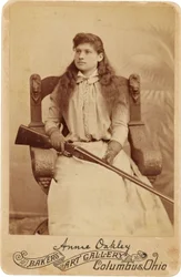 Annie Oakley : Annie Oakley:. Aquí, la legendaria tiradora está sentada con bastante recato en una enorme silla de estilo renacentista, con una escopeta en el regazo. Sus delicados rasgos femeninos y su cabello suelto desmienten su robusta imagen al aire l