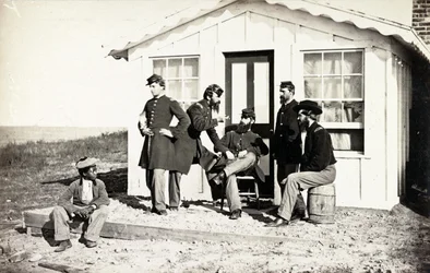 Cinco soldados de la Guerra Civil reunidos en el porche de tierra fuera de una casa, joven afroamericano sentado cerca de ellos, 1861-65