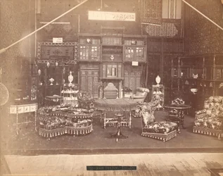 Exhibición de S. Howard & Sons -- Edificio Principal, Colección de muebles, incluyendo sillas tapizadas, mesas, una chimenea, gabinetes y estanterías. En las paredes hay alfombras, y en las mesas y estantes hay jarrones