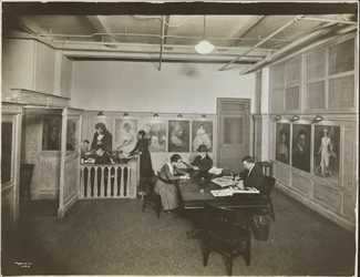Oficina principal/sala de recepción en la Mutual Film Co. con secretaria en el escritorio, personas en una mesa leyendo y retratos en la pared, Nueva York