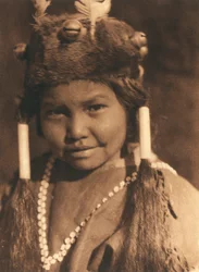 Primer plano de un niño indio americano de la tribu o aldea Klamath. Lleva un gorro de piel con bellotas y plumas aplicadas y el pelo en dos coletas