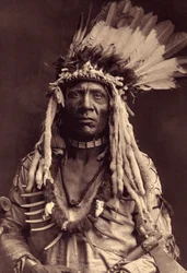 Cola de Comadreja (Apohsuyis). Este indio piegan lleva el famoso seto de guerra de plumas de águila y piel de comadreja, una túnica de ante, dos collares, uno de hueso y otro de garras de oso pardo, y una pipa tomahawk. Foto tomada del volumen 6 de la enci