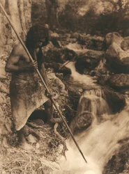 Joven indio americano de la tribu o pueblo Hupa pescando salmón con un palo largo. Lleva una piel de animal atada al lomo. Detrás de él, un bosque y un arroyo.