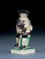 Jarra de Dick Whittington, moldeada levantándose de una silla de respaldo alto, sosteniendo a su gato, salpicada en ropa marrón, verde y negra, un escudo moldeado a su lado