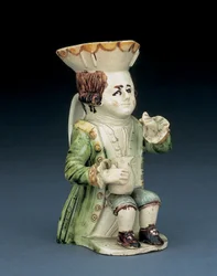 Jarra Toby del bebedor, su sombrero con borde festoneado marrón y amarillo, su cabello castaño inciso recogido en coleta y con una roseta blanca, sentado con una jarra abalaustrada sobre la rodilla, ca. 1770