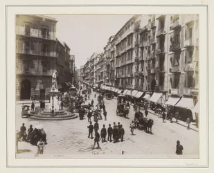 Vista de la Via Roma de Nápoles con una estatua, tiendas, carruajes tirados por caballos y transeúntes