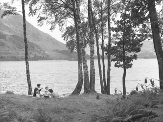 Una familia descansa cerca de un lago con árboles a la derecha y montañas al fondo.