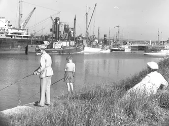 Un hombre y un niño pescando en Glasson Dock mientras una dama sentada detrás observa, varios barcos están amarrados