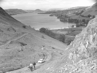 Vista de una ladera con carretera, coches y gente, lago al fondo.