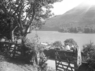 Cuatro personas sentadas en un muro de piedra seca haciendo un picnic con vistas al lago, el coche aparcado con el maletero abierto