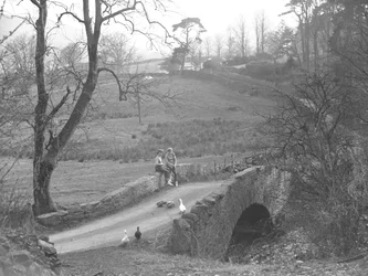 Dos niñas sentadas en un puente de piedra sobre el río Cocker al pie de Scale Hill, observando gansos en el camino de Loweswater