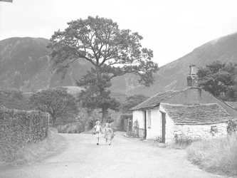 Dos chicas jóvenes de la mano sosteniendo ramitas de dedaleras caminando por la carretera pasando por delante de la casa de campo hacia la cámara.