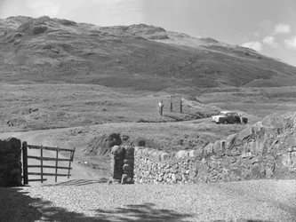 Vista de la carretera que pasa por Hard Knott Pass, dos personas de pie cerca de una señal de tráfico y un coche aparcado en el arcén de hierba.