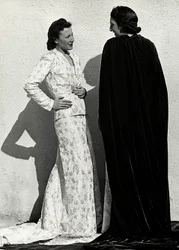 Dos modelos presentan creaciones de la casa de alta costura italiana "Dragoni". Una modelo lleva un conjunto de material estampado compuesto por una chaqueta y una falda larga con cola. La otra modelo, vista desde atrás, lleva un manto largo de terciopelo.