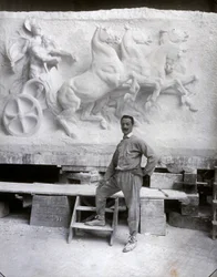 Retrato de cuerpo entero del famoso escultor Pietro Canonica. Está posando en su estudio frente a un alto relieve de mármol en el que está trabajando
