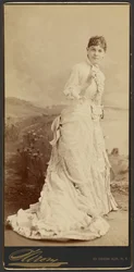 [Mujer no identificada con un largo y elegante vestido blanco, de pie]