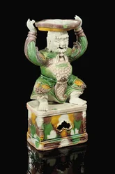 Candelero de espiga vidriado en amarillo huevo y verde espinaca, modelado como un inmortal sonriente con barba ondulante y los brazos levantados para sostener el accesorio cuadrilobulado con espiga, equilibrado sobre su cabeza, Kangxi