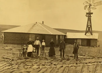 Familia de colonos ante su casa de césped, con un molino de viento en el tejado del edificio contiguo, en el condado de Custer, Nebraska, ca. 1885
