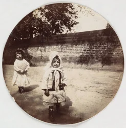 Dos niños pequeños caminando junto a una pared, c 1890