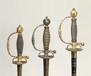 Empuñaduras de tres espadas francesas: una de acero, azulada y sobrepuesta con diseños en oro cincelado, c.1730; una de acero perforado y cincelado y el fondo de oro mate, c.1760; una de acero cincelado
