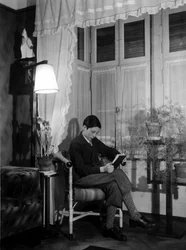 Un adolescente sentado en un interior burgués lee un libro, lleva pantalones cortos y calcetines altos. 1915. Foto histórica, mujer leyendo, ca. 1915