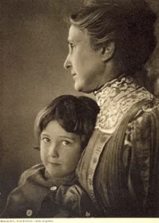 Retrato de medio cuerpo de un niño tiernamente abrazado por una anciana. La mujer posa de perfil, sosteniendo el pecho del pequeño. Tiene una mirada vivaz y pícara, que dirige hacia la cámara.