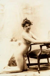 Retrato desnudo de una mujer joven arrodillada delante de un pequeño sillón.