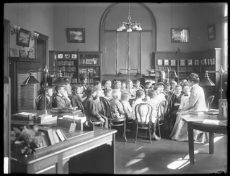 Niños escuchan a una mujer en la sala de lectura de una sucursal no identificada de la Biblioteca Pública del Borough de Queens, c.1910