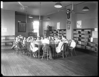 Niños escuchan a una mujer en la sala de lectura de una sucursal no identificada de la Queens Borough Public Library, c. 1910