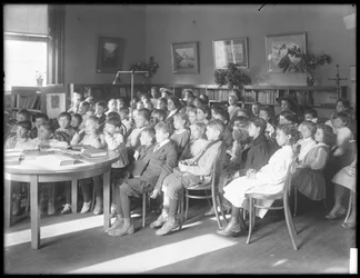 Niños sentados, escuchando en la sala de lectura de una sucursal no identificada de la Biblioteca Pública del Distrito de Queens, c.1910