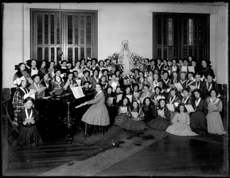 Retrato de grupo de una clase de coro de niñas reunidas alrededor de un piano con partituras, probablemente en el Asilo de Huérfanos Católico Romano en Kingsbridge, Bronx, c.1911
