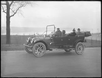 Tres personas no identificadas y dos perros Chow conduciendo en un coche convertible a lo largo de un río o lago, c.1912
