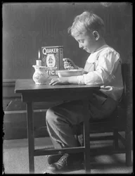 William Gray Hassler sentado en una pequeña mesa comiendo un tazón de cereal de arroz inflado Quaker, c.1912